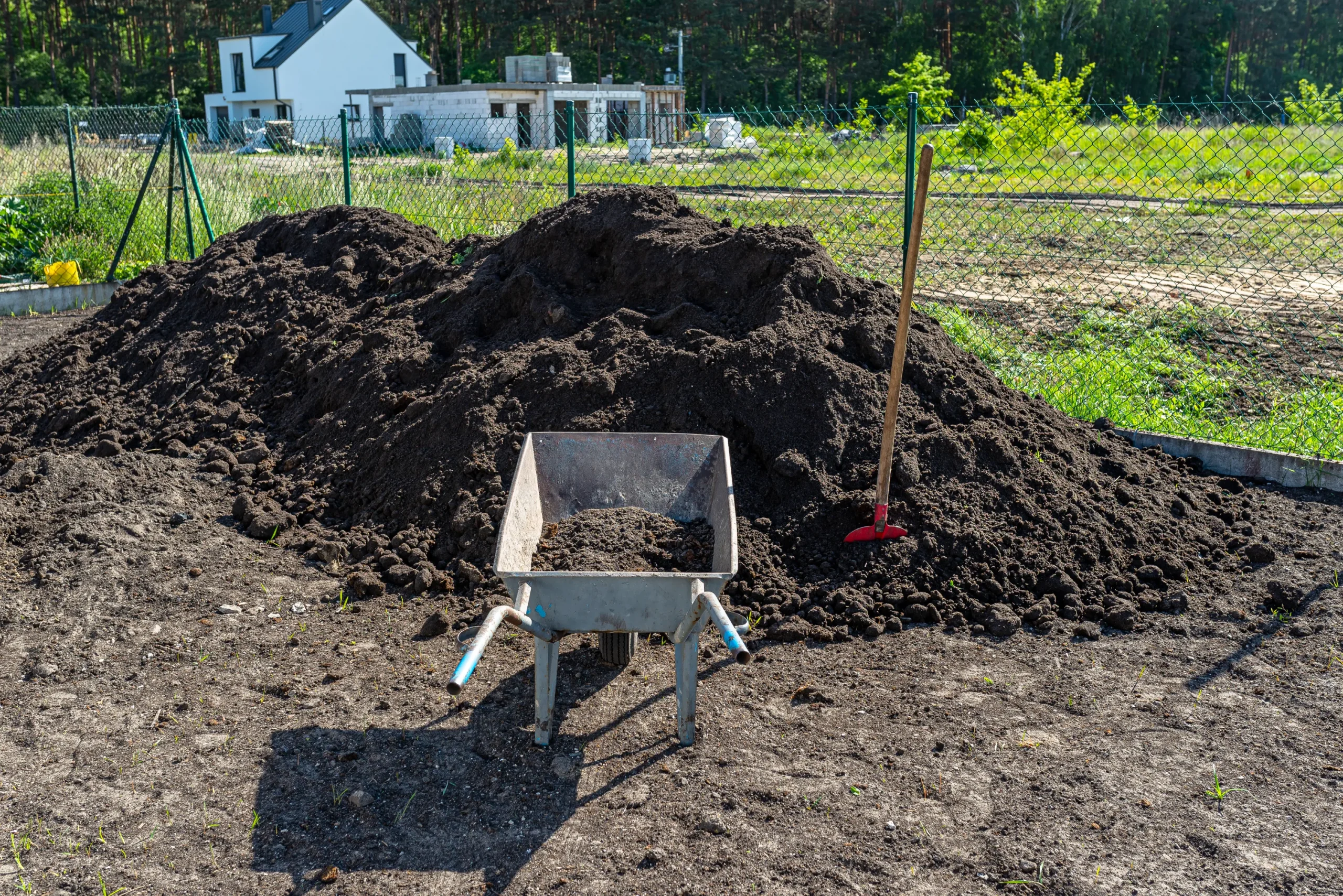 A large pile of dark soil with a wheelbarrow and shovel in front, situated in a sunny garden area. A modern white house is visible in the background.