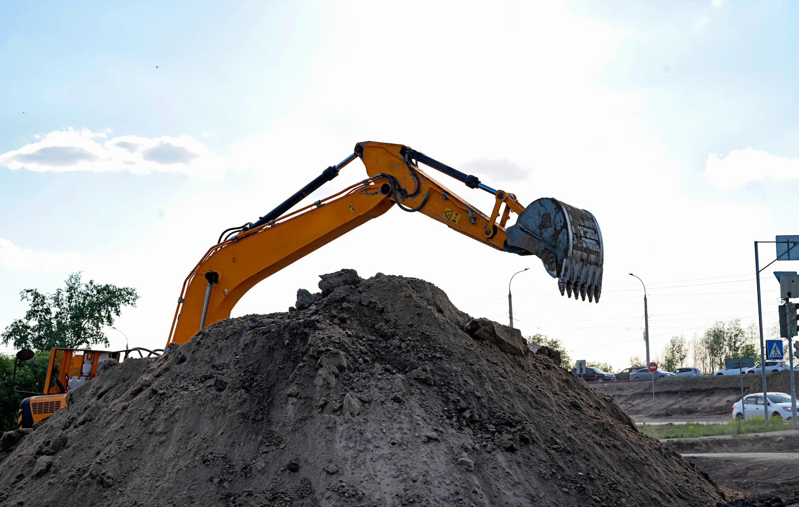 An orange excavator with a raised arm and bucket is working on a large dirt pile under a bright sky. Cars and trees are visible in the background.