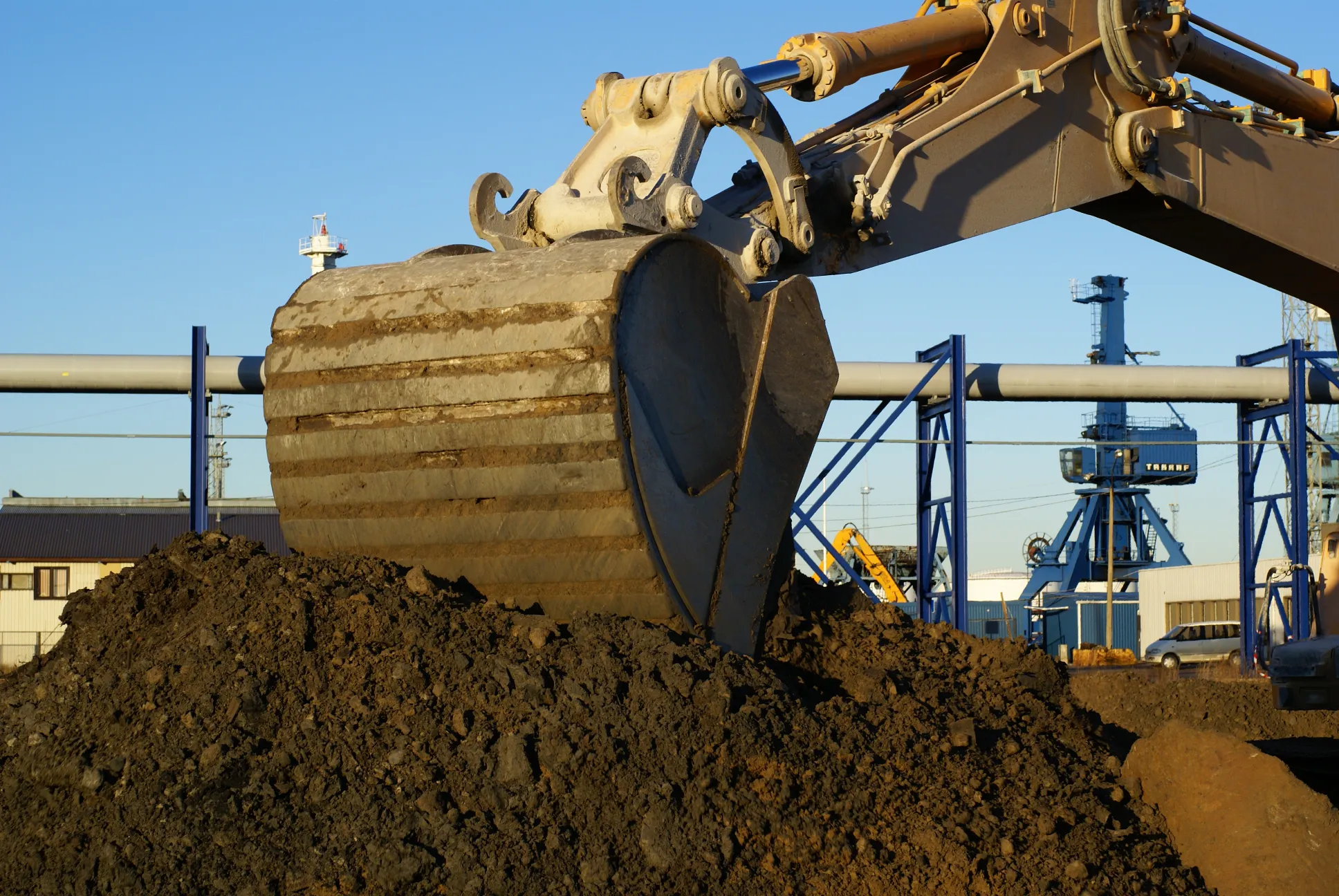 Excavator bucket digging into a large dirt pile under a clear blue sky, with industrial structures and cranes visible in the background.