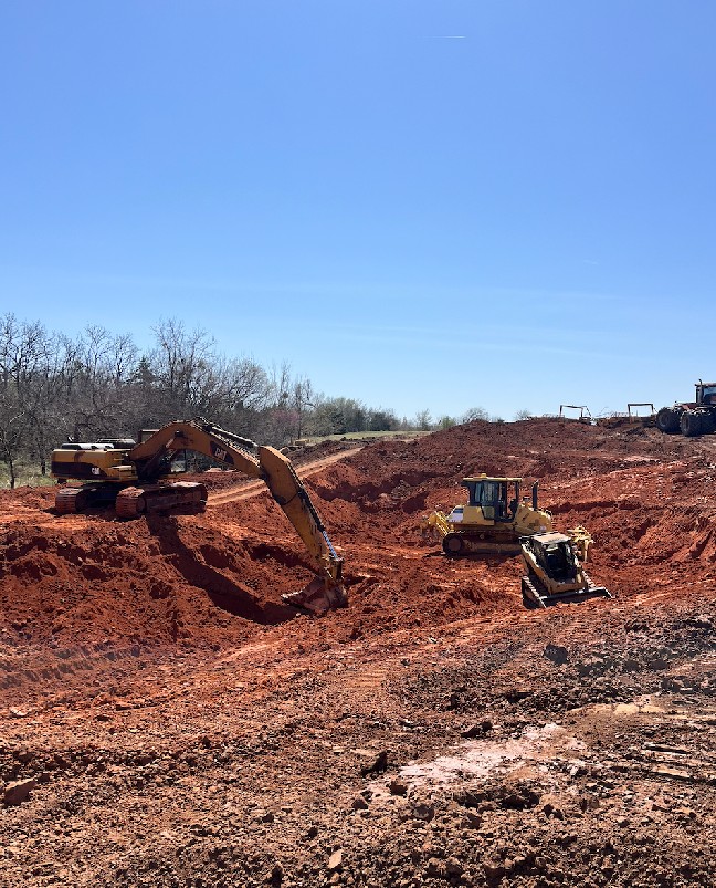 Three yellow construction vehicles, including excavators and a bulldozer, work on red earth under a clear blue sky in a rural area. Sparse trees line the background.