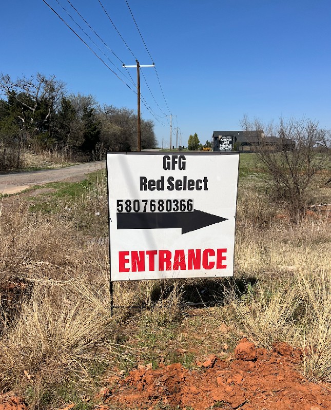 Sign on grassy roadside under clear blue sky reads, "GFG Red Select" and "5807680366," with an arrow pointing right and "ENTRANCE" in red.