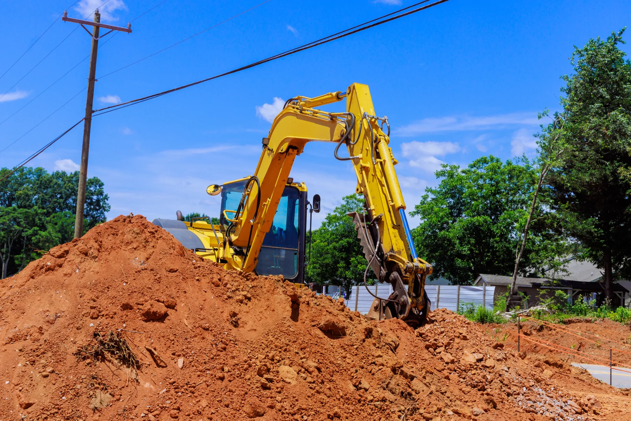 A yellow excavator sits on a mound of red dirt at a construction site, surrounded by trees under a bright blue sky with minimal clouds.