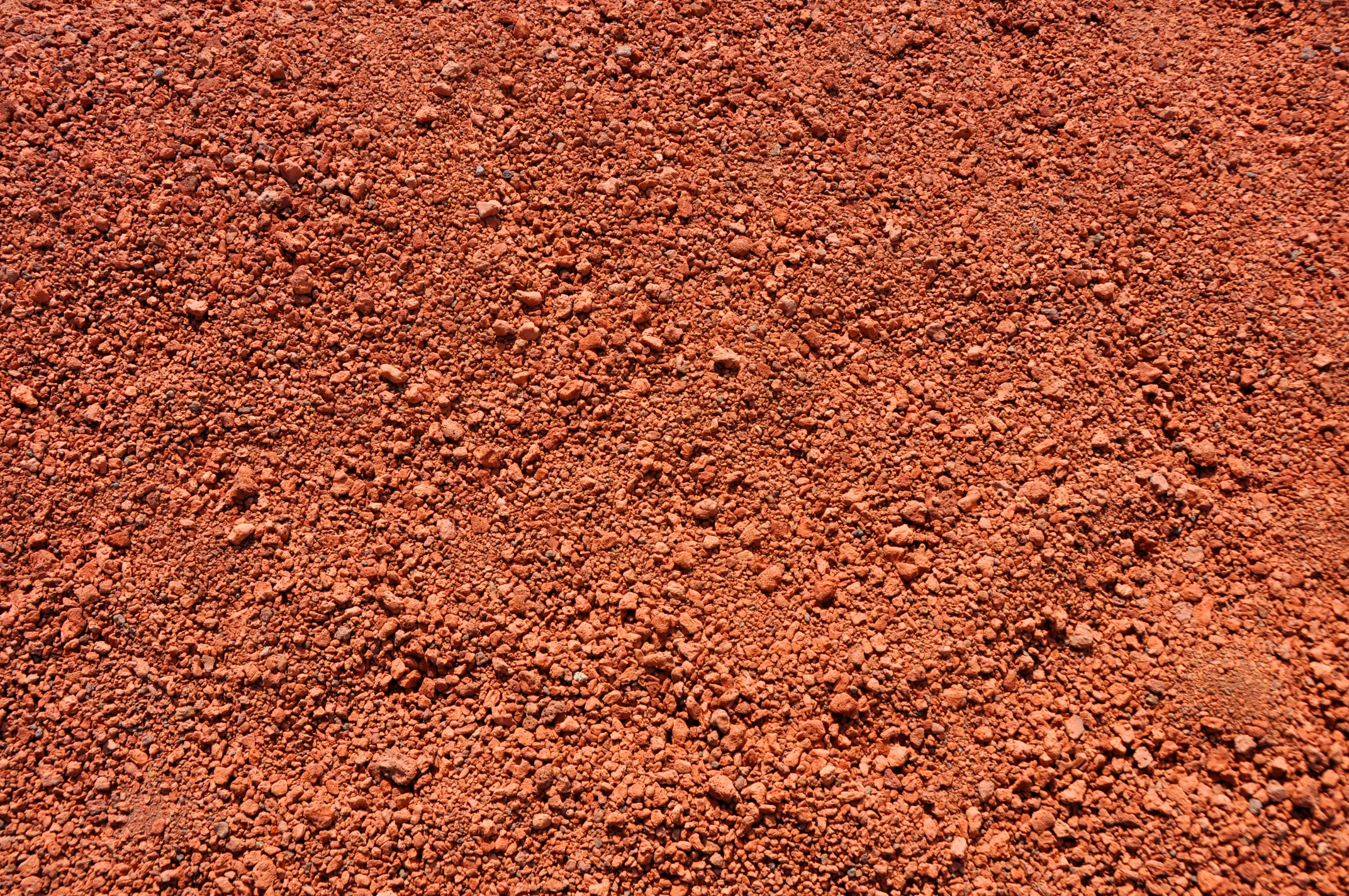 Close-up of a textured red dirt surface with small pebbles and grains, conveying a rough, earthy feel. Warm tones dominate the image.