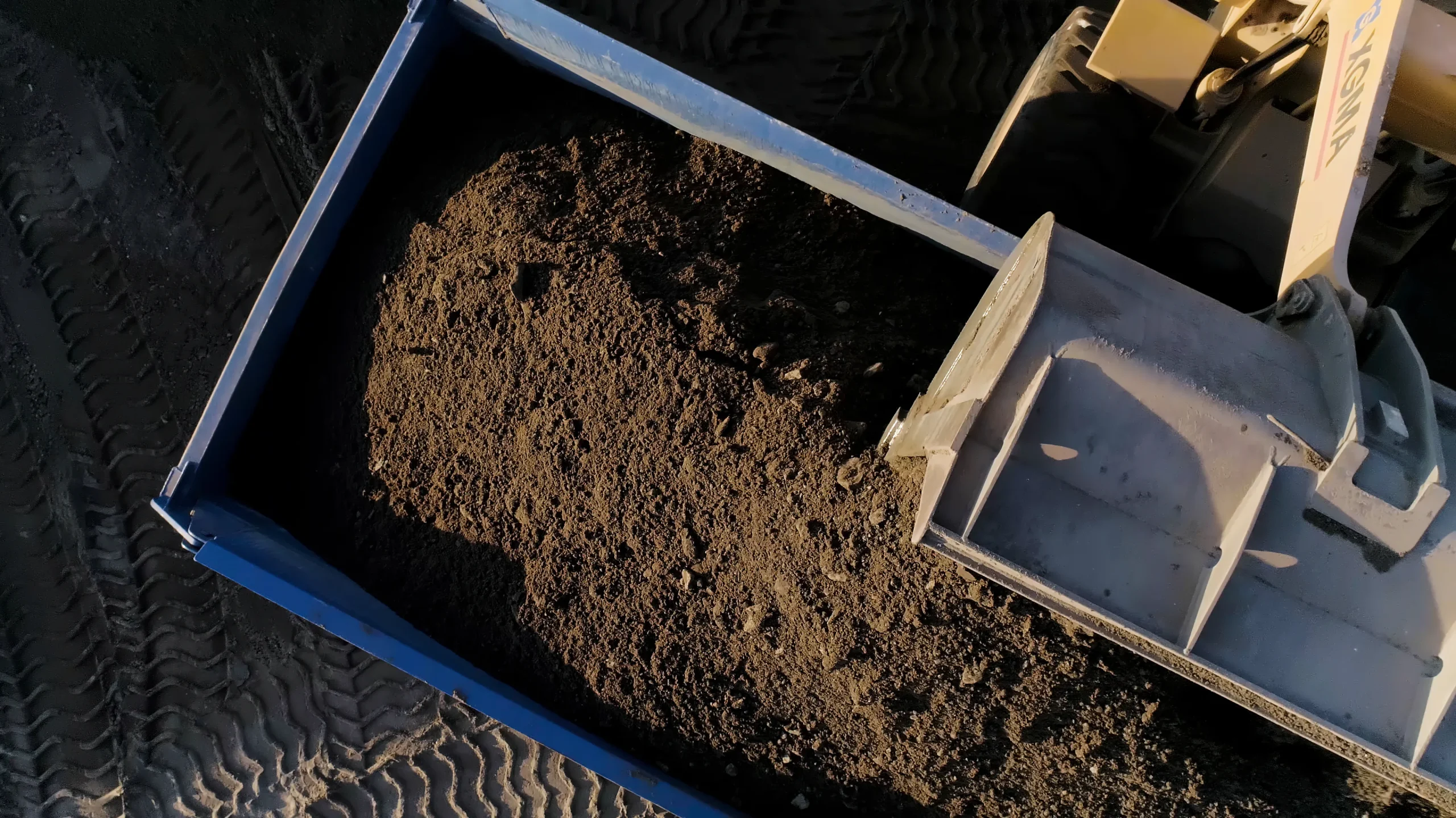 Overhead view of a dump truck bed being filled with dark soil by a loader. Sunlit earth and textured tire tracks create a rugged, industrious scene.
