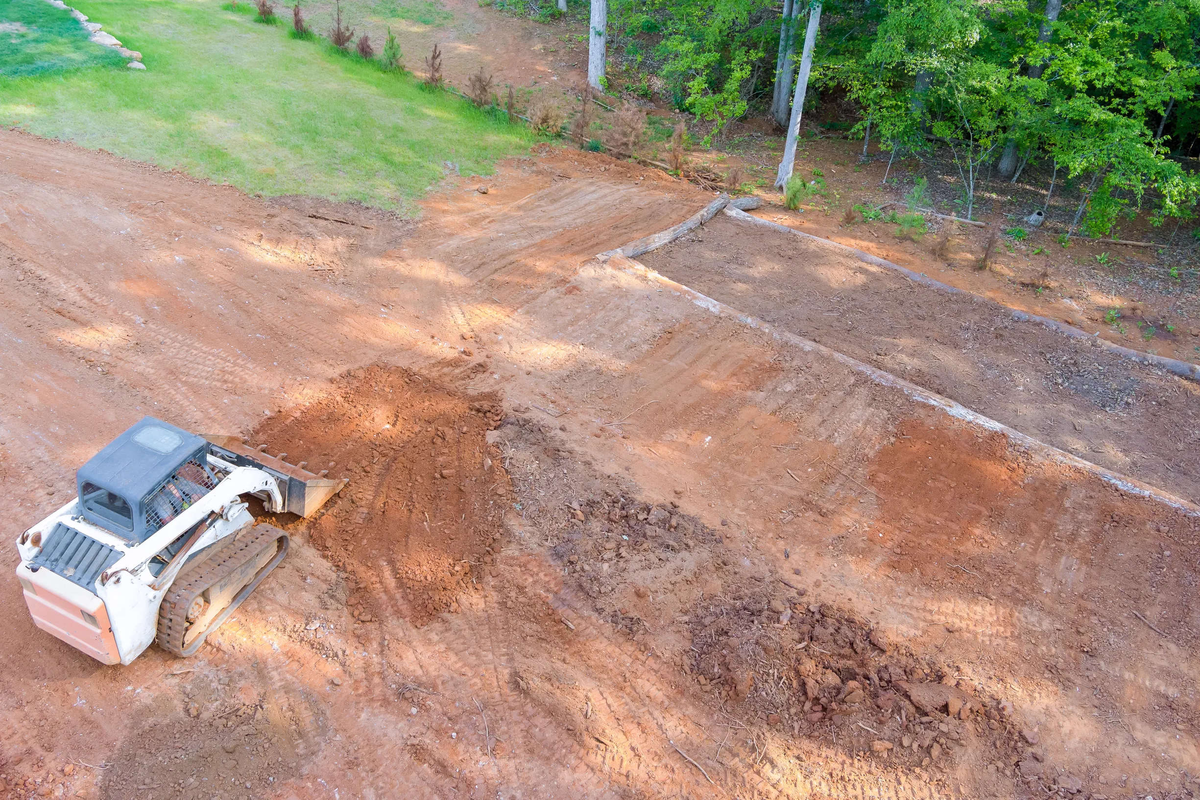 A white bulldozer is leveling a large, reddish-brown dirt area near a patch of green grass. Trees line the background, suggesting a construction site.